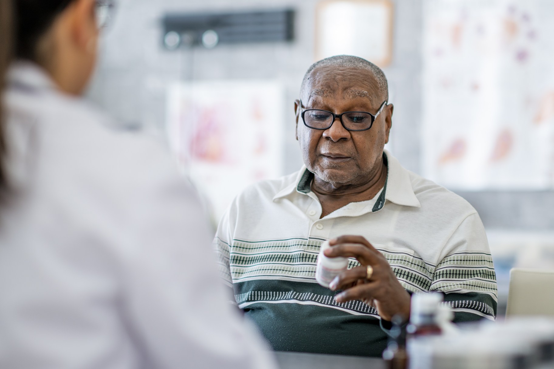 Un adulto mayor sostiene y observa un frasco de píldoras mientras está sentado frente a un médico en el consultorio del médico. Parece estar tomando una decisión al respecto.