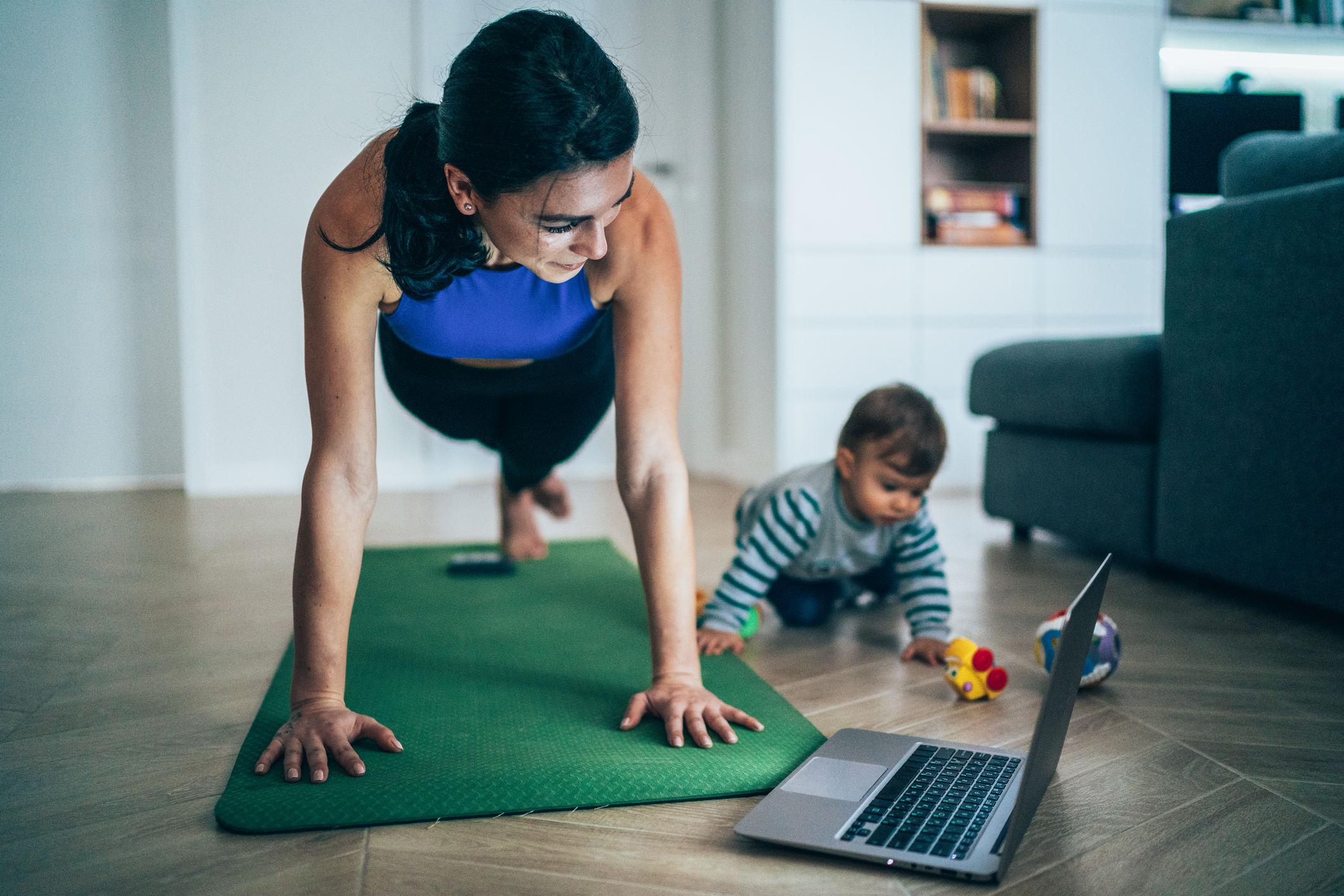 Mujer se ejercita mientras ve videos de ejercicios en una computadora portátil y su bebé juega cerca