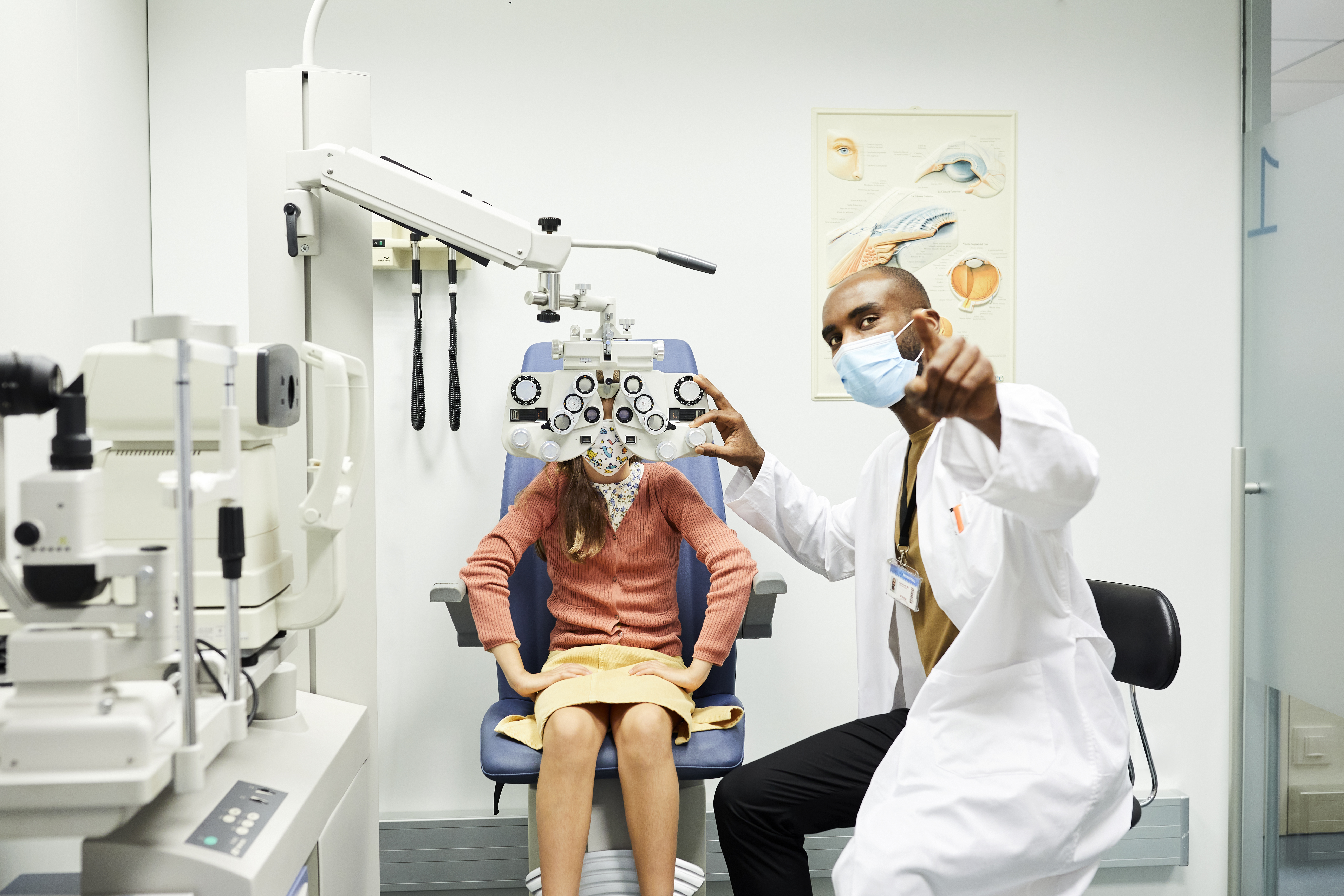 Oftalmólogo señalando a una niña mientras usa una mascarilla protectora. La niña está sentada detrás de la optometría en el consultorio del médico. Están en el hospital para un examen de la vista durante la crisis del COVID-19.