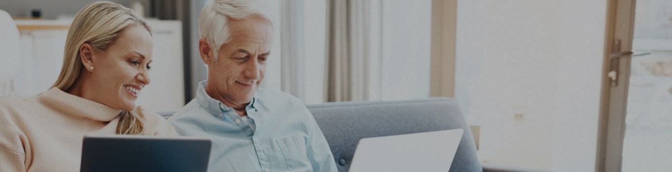 Mujer rubia con una computadora portátil sentada en un sofá junto a un hombre mayor con una computadora portátil.