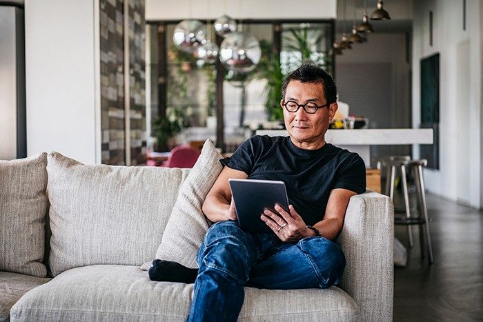 Hombre chino de unos 50 años relajándose en casa sobre el sofá, ropa casual, leyendo libros electrónicos, navegando por internet